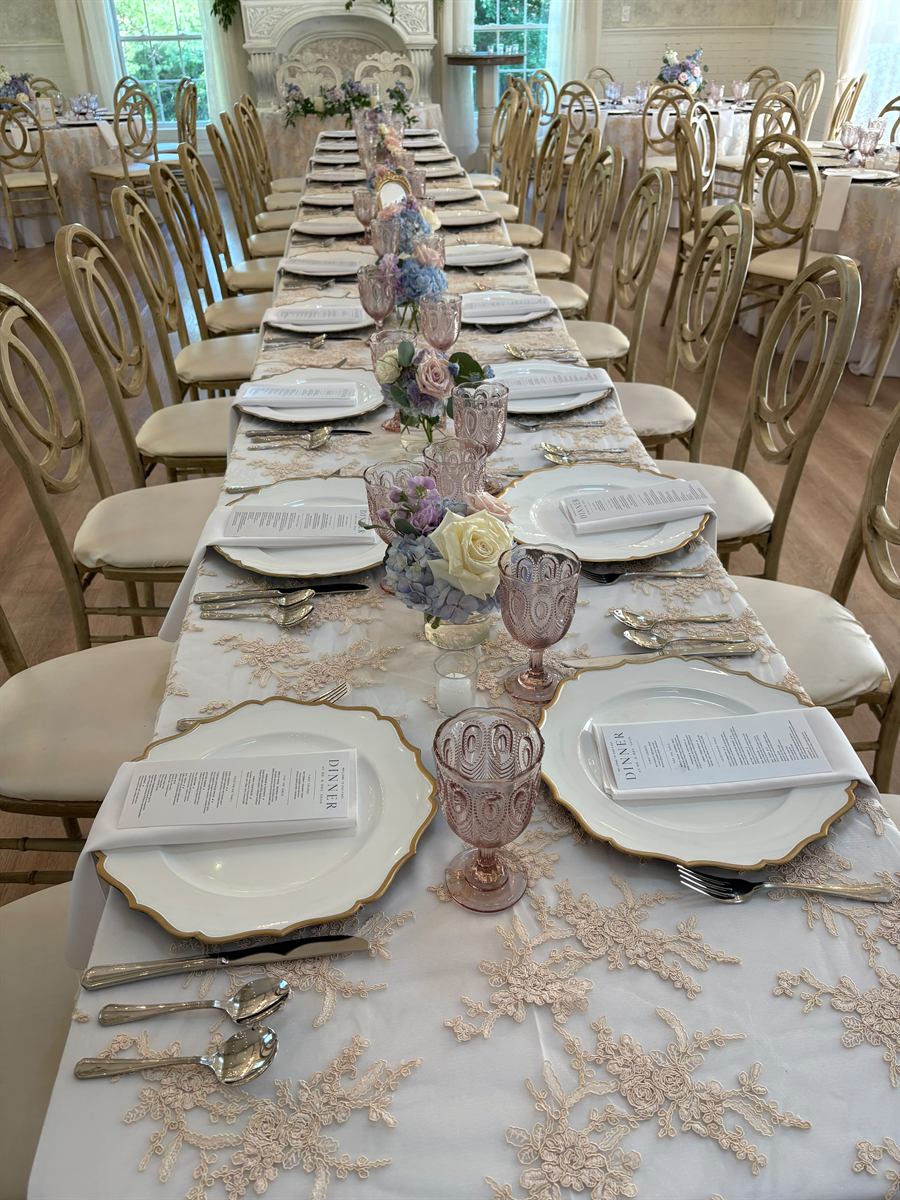 A long banquet table set end to end with gold-rimmed chargers, blush lace linen and pink glassware