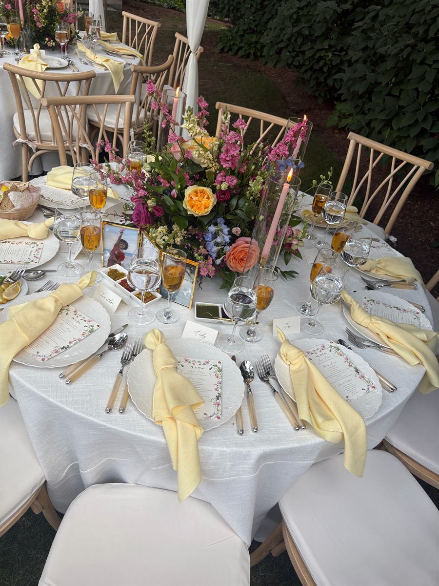An outdoor table at dusk with yellow linen napkins, taper candles and a bright floral centrepiece