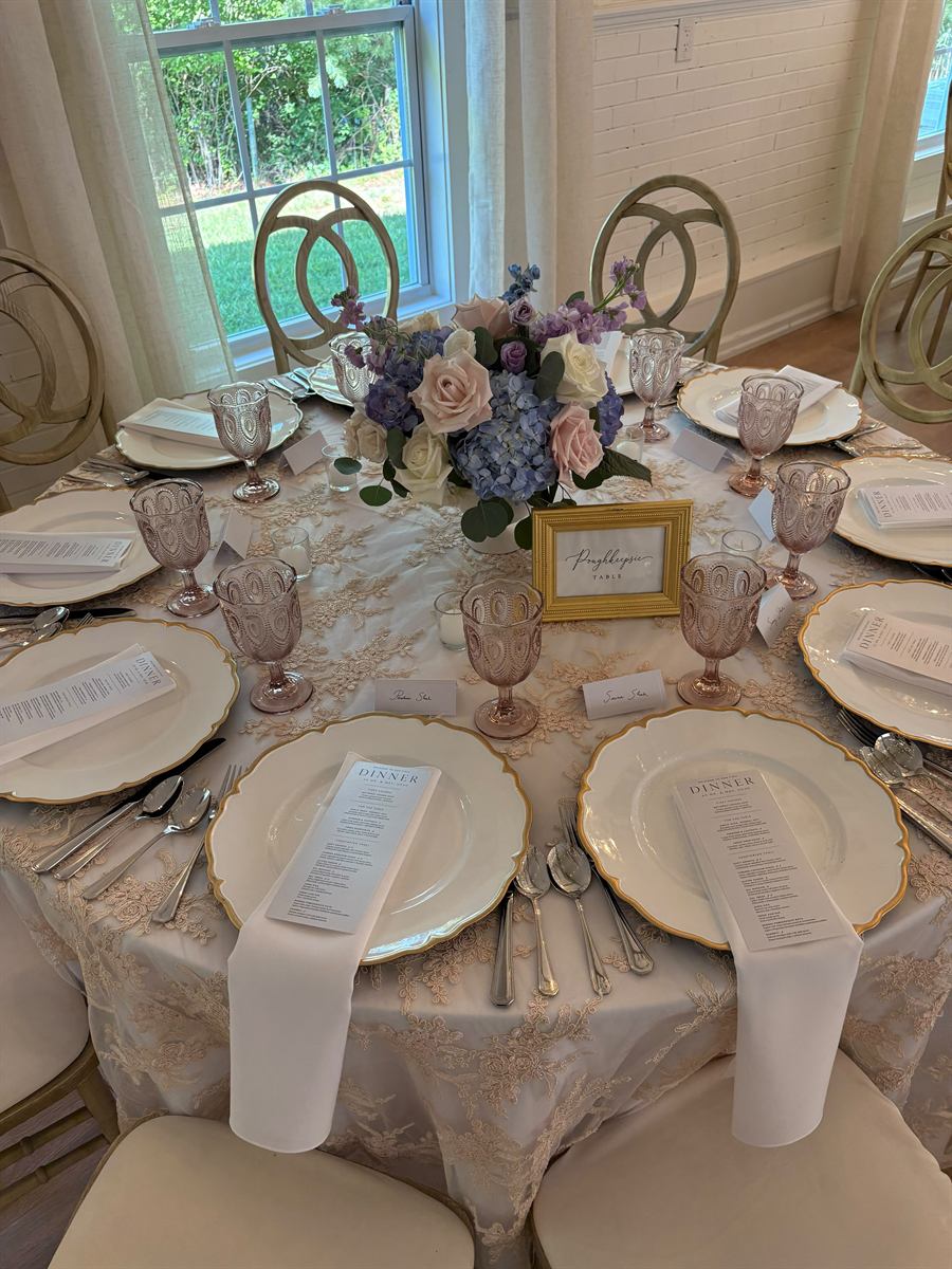 A round table dressed in blush lace with gold-rimmed chargers, pink goblets and a blue and lavender centrepiece